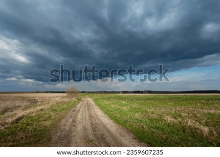 Unpaved road through meadows and dark cloud on the sky, spring day