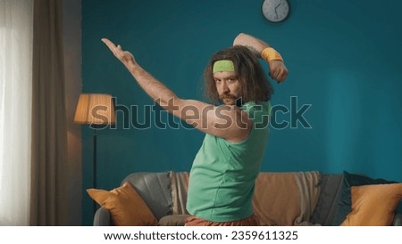 A middle aged man with long hair and beard is coached in the apartment. The athlete poses and shows his muscles with hand movements. Fitness at home. Medium shot. Royalty-Free Stock Photo #2359611325