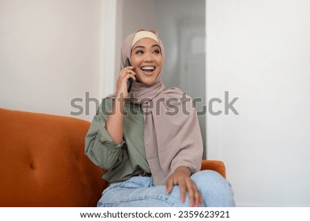 Mobile Communication. Joyful Black Lady In Hijab Talking On Cellphone Sitting On Sofa At Home, Enjoying Conversation, Communicating With Friends Via Smartphone, Looking Aside