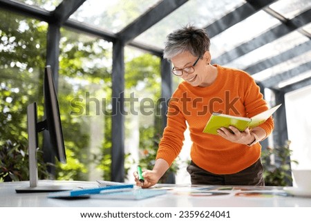 Stylish middle aged woman designer working with color palette and computer pc in home office, architect choosing colors for decoration, taking notes in notepad, coworking space, free space