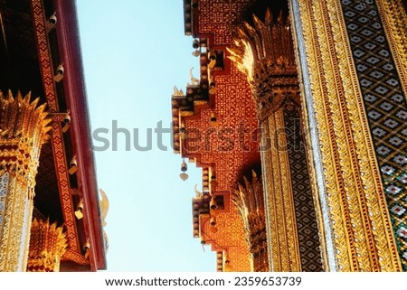 The image portrays a temple in Thailand, adorned with intricate gold and red patterns. Small statues embellish the roof. The photo, taken from a low angle, offers a view of the sky in the background.