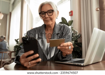 confident well-groomed senior woman with gray hair and glasses dressed in a gray jacket sits in a cafe and holds out a smartphone and a credit card to pay the bill