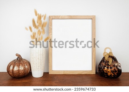 Mock up portrait wooden frame with fall grasses and pumpkin decor on a wood shelf against a white wall. Autumn concept. Copy space.