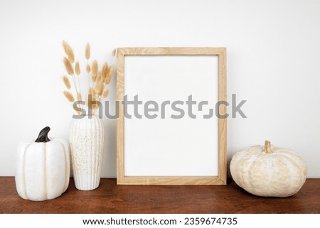 Mock up portrait wooden frame with fall grasses and pumpkin decor on a wood shelf against a white wall. Autumn concept. Copy space.
