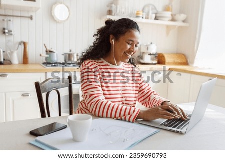 Side view of happy african american female freelancer working on laptop sitting at kitchen table, making notes in her diary, chatting online in corporate chat or messenger, wearing wired earphones