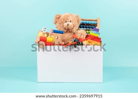 Toy box full of baby kid toys. Container with teddy bear, fluffy and educational wooden toys on light blue background. Cute toys collection for small children. Front view Royalty-Free Stock Photo #2359697915