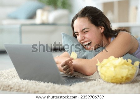 Funny woman eating potato chips watching media on laptop lying on the floor laughing hilariously at home Royalty-Free Stock Photo #2359706739