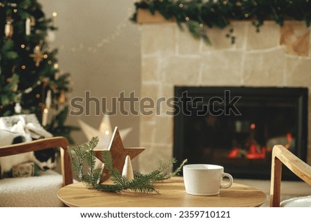 Stylish white cup with wooden star decor on table against stylish festive christmas tree with golden lights and cozy fireplace. Cup mock up, christmas advertising product template. Space for text