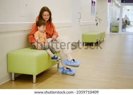 A patient and her baby sit in the clinic corridor, watching videos on their phone while waiting for their turn to see the doctor. Kid boy aged two years (two-year-old)