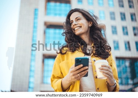 Beautiful young woman is using an app in her smartphone device to send a text message near business buildings. Young positive businesswoman with take away coffee cup in hand using mobile phone 