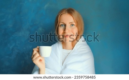 Happy young woman waking up after sleeping wrapped in white soft comfortable blanket holds cup of coffee in morning on blue background at home