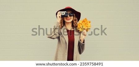 Autumn color style outfit, beautiful elegant woman photographer taking picture with film camera holds yellow maple leaves wearing hat on gray background