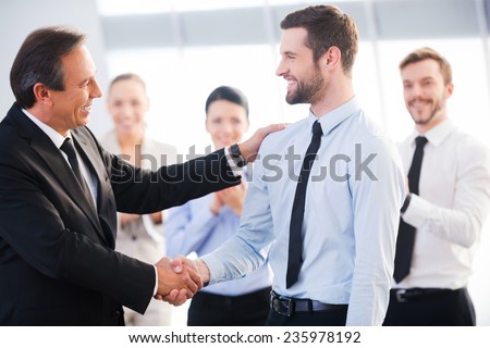 Good job! Two cheerful business men shaking hands while their colleagues applauding and smiling in the background Royalty-Free Stock Photo #235978192