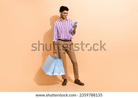 Full size photo of clever man dressed striped shirt look at smartphon make online order hold new outfit isolated on beige color background
