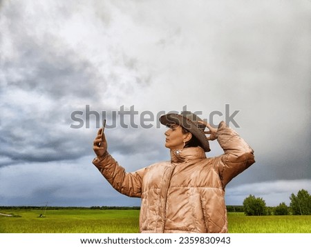 An adult girl in a field and with a stormy sky with clouds takes pictures of a rainbow and takes a selfie in the rain. A woman having fun outdoors on rural and rustic nature