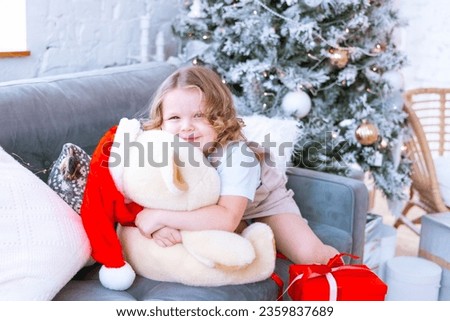 cute happy little girl with curls in Santa hat playing with toys teddy bear and new gifts near Christmas tree at home, New Year and Christmas family concept