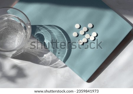 Minimal medicine concept, White pills on blue notebook, glass with water on white table background with sunlight shadows. Royalty-Free Stock Photo #2359841973