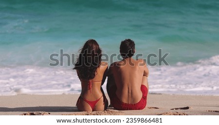 Man and woman talking while sitting by the ocean on a sandy beach. A brunette Caucasian woman and a man are sitting with their backs to the camera, looking at the waves. The concept of a romantic