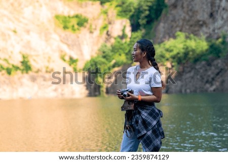 young adventurer standing on the shore of a lake and posing while holding an analog camera. Beautiful woman posing for a picture on her holiday