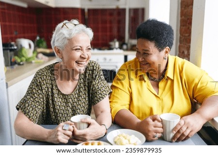 Two senior female friends of diverse ethnicity laughing together at funny situation in past or jokes drinking coffee with sweats at kitchen table, recollecting best memories of their lives Royalty-Free Stock Photo #2359879935