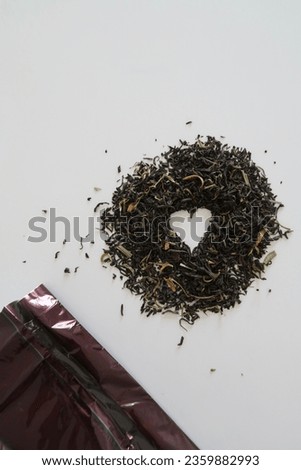 Pile of black tea leaves next to a brown closed bag of tea, shape of heart in the pile of tea leaves, natural light, neutral tones, no people, closeup image of dry black tea leafs, detail picture
