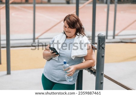 A plus size female, post workout, enjoys her smartphone outdoors. She beams with happiness and satisfaction, a water bottle in hand. Royalty-Free Stock Photo #2359887461