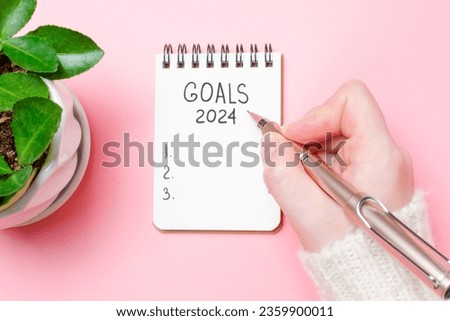 Female hand writes in a paper notebook goals 2024 close-up on a pink background. Royalty-Free Stock Photo #2359900011