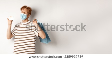 Concept of covid-19 and lifestyle. Happy young shopper in face mask holding shopping bag and showing plastic credit card, buying with discounts, white background.