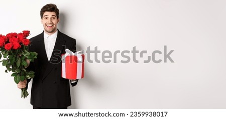 Concept of holidays, relationship and celebration. Image of handsome smiling guy in black suit, holding bouquet of red roses and giving present on new year, white background.