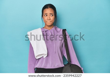 Filipina with gym gear on blue studio confused, feels doubtful and unsure. Royalty-Free Stock Photo #2359942227