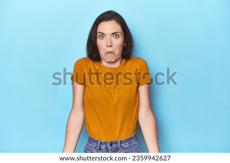 Young caucasian woman on blue backdrop shrugs shoulders and open eyes confused.