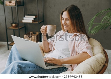 Young IT woman wear casual clothes sits in armchair hold use work on laptop pc computer drink tea stay home hotel flat rest relax spend free spare time in living room indoor. Lifestyle lounge concept Royalty-Free Stock Photo #2359943455