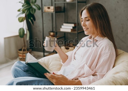Side view young happy calm woman wear casual clothes sits in armchair read book drink coffee tea stay at home hotel flat rest relax spend free spare time in living room indoor Lifestyle lounge concept Royalty-Free Stock Photo #2359943461