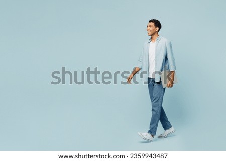 Full body side view young man of African American ethnicity wear shirt casual clothes hold closed laptop pc computer walking going solated on plain pastel light blue cyan background studio portrait