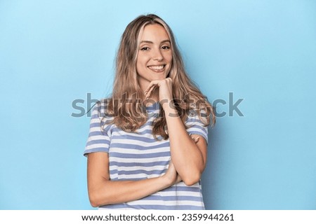 Blonde young caucasian woman in blue studio smiling happy and confident, touching chin with hand.