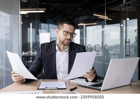 A worried young man businessman, accountant reviews documents, business agreements, bills in the office. He sits at the desk in the office and looks at the papers. Royalty-Free Stock Photo #2359958203