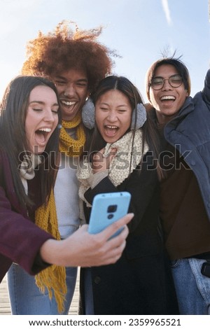 Vertical group excited friends taking selfie mobile photo looking at screen phone in warm clothes. Young surprised multiracial people having fun in cold and sunny winter day outdoor. Generation z.