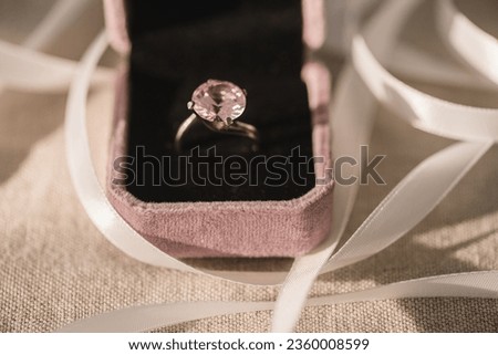 A closeup of a pink diamond ring in a velvet box and ribbon on a burlap mat
