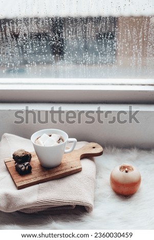 Winter food and drink on folded sweater nearby window with rain drops and candle on fur rug.  Royalty-Free Stock Photo #2360030459