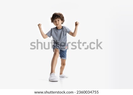 Strongman. One curly Caucasian preschool boy in casual clothing with hands up posing isolated on white studio background. Copyspace for ad Royalty-Free Stock Photo #2360034755