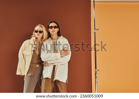 fall fashion and trends, interracial women in sunglasses and outerwear posing on duo color backdrop Royalty-Free Stock Photo #2360051009