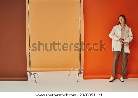 brunette asian woman in autumn attire standing on multicolored backdrop, leather pants and outerwear Royalty-Free Stock Photo #2360051121