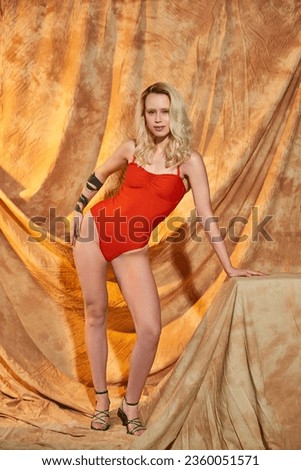 tattooed woman in orange swimwear posing on beige backdrop with drapery, blonde model in studio