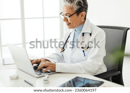 Senior doctor using a laptop to access medical software, which allows her to practice efficiently. Female physician working with technology in her office, wearing a white lab coat and a stethoscope. Royalty-Free Stock Photo #2360064197