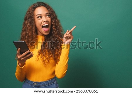 Beautiful young woman points thumb to copy space left, cellphone, green studio Royalty-Free Stock Photo #2360078061