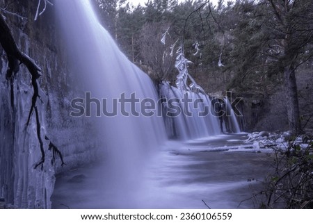 Frozen waterfall in a natural and scenic environment in the mountains of Madrid. Pradillo reservoir in winter with the water half frozen after the last snowfalls. Frozen waterfall in long exposure.  Royalty-Free Stock Photo #2360106559
