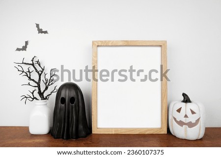 Halloween mock up. Wooden frame on a wood shelf with black branches, ghost and jack o lantern decor. Portrait frame against a white wall. Copy space.