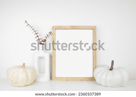 Mock up portrait wooden frame with fall braches and pumpkin decor on a white shelf against a white wall. Autumn concept. Copy space.