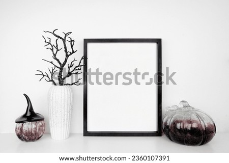 Halloween mock up. Black frame on a white shelf with black branches and glass pumpkin decor. Portrait frame against a white wall. Copy space.