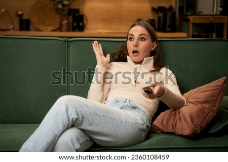 A woman holds a TV remote control and covers her eyes with her hand while sitting on a green sofa in the living room. A young woman switches the TV channel in fear. 